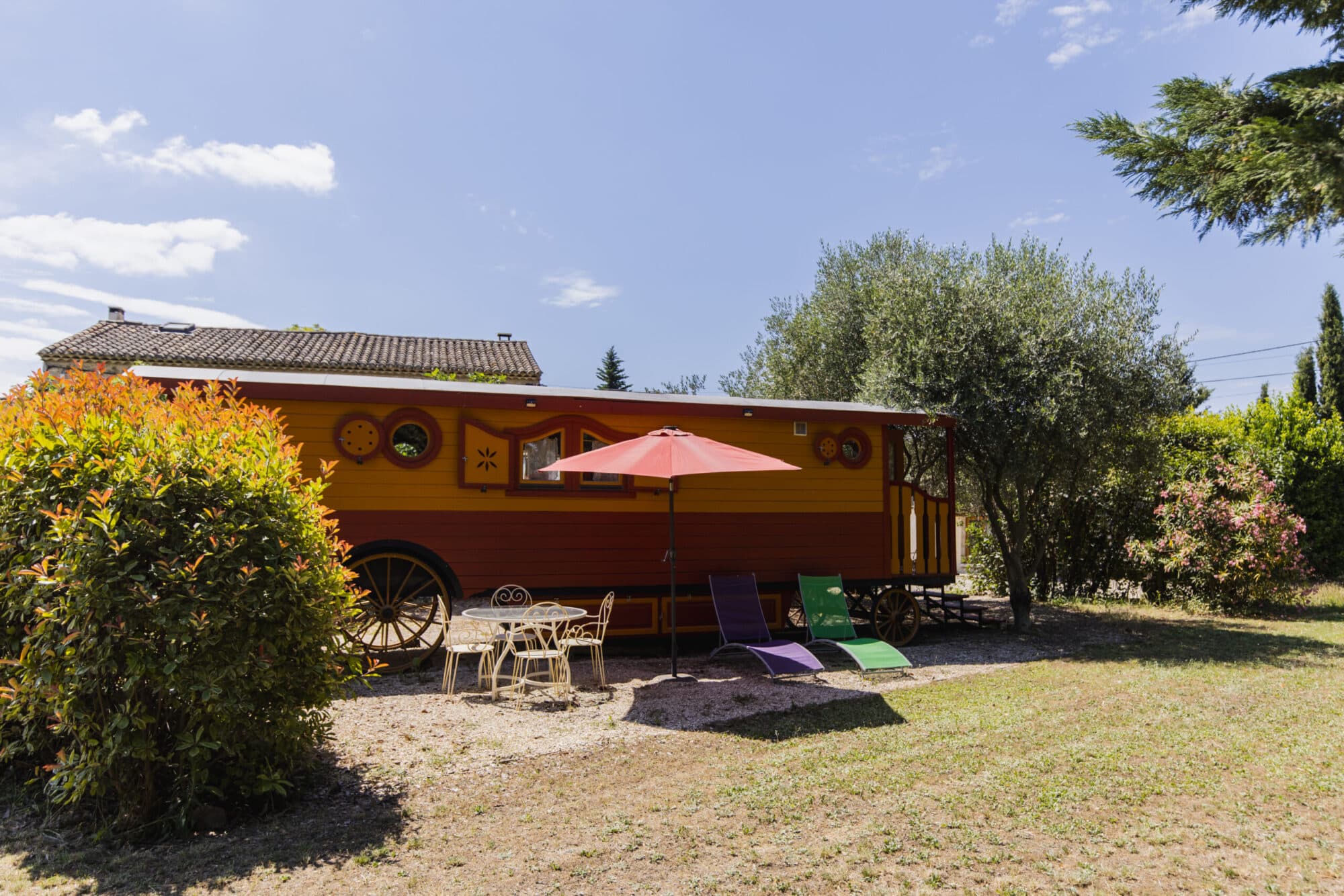Roulotte jaune et rouge dans le jardin, avec parasol, table en fer forgé, deux transats et oliviers, en Luberon