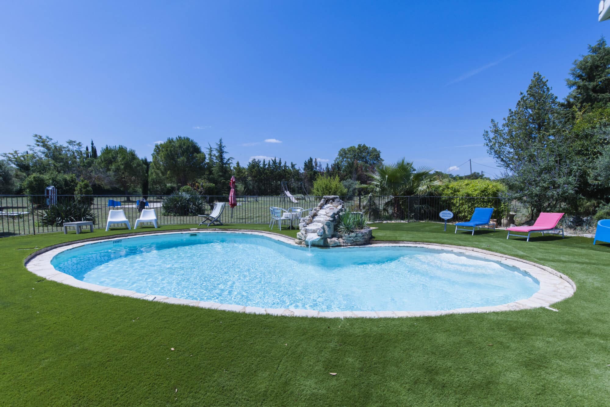 Piscine à la forme arrondie avec cascade de rochers, entourée de pelouse, de transats et d’arbres, en Luberon