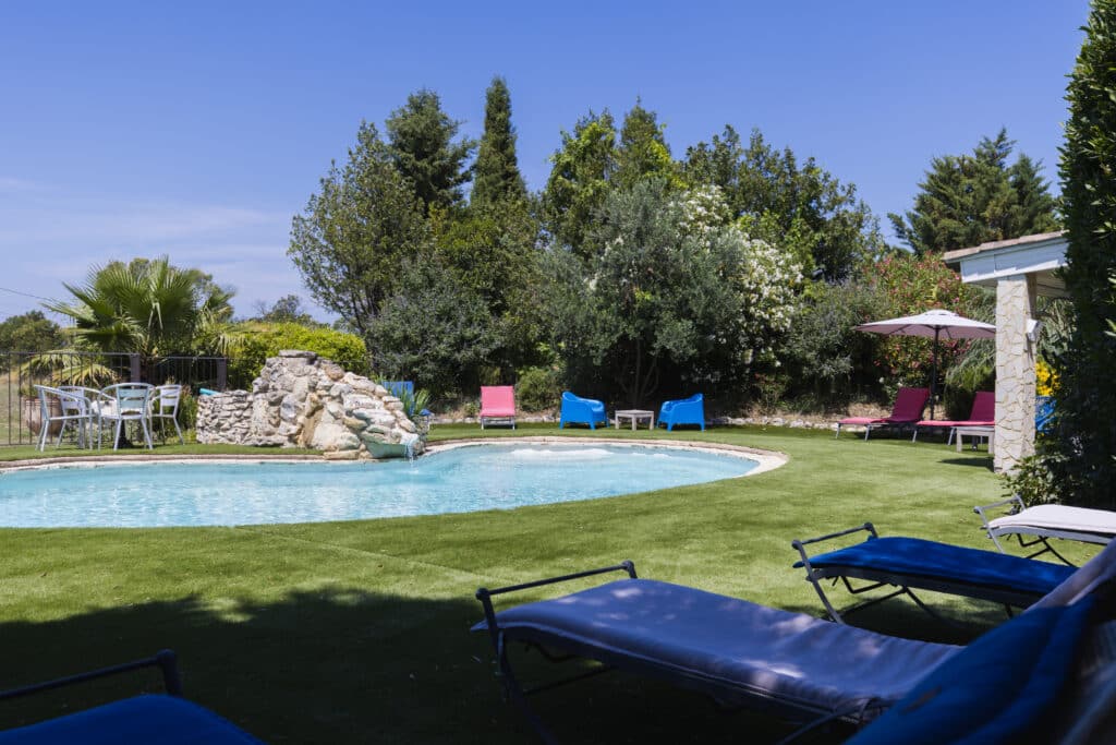 Piscine avec cascade de rochers entourée de pelouse, avec bains de soleil bleus au premier plan, transats roses, parasol et salon de jardin au fond
