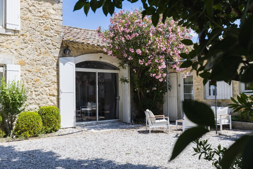 Entrée en arcade avec baie vitrée et volets blancs, laurier-rose en fleurs, buis taillés et fauteuils de jardin blancs sur le gravier