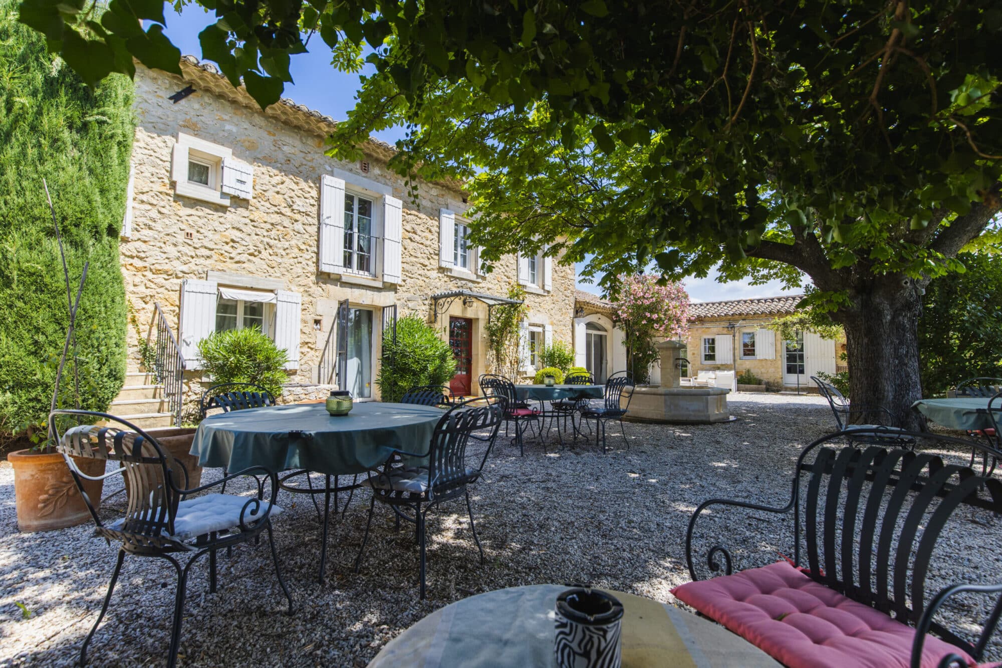 Façade en pierre du mas avec volets blancs, table dressée sur la terrasse et grand arbre ombragé