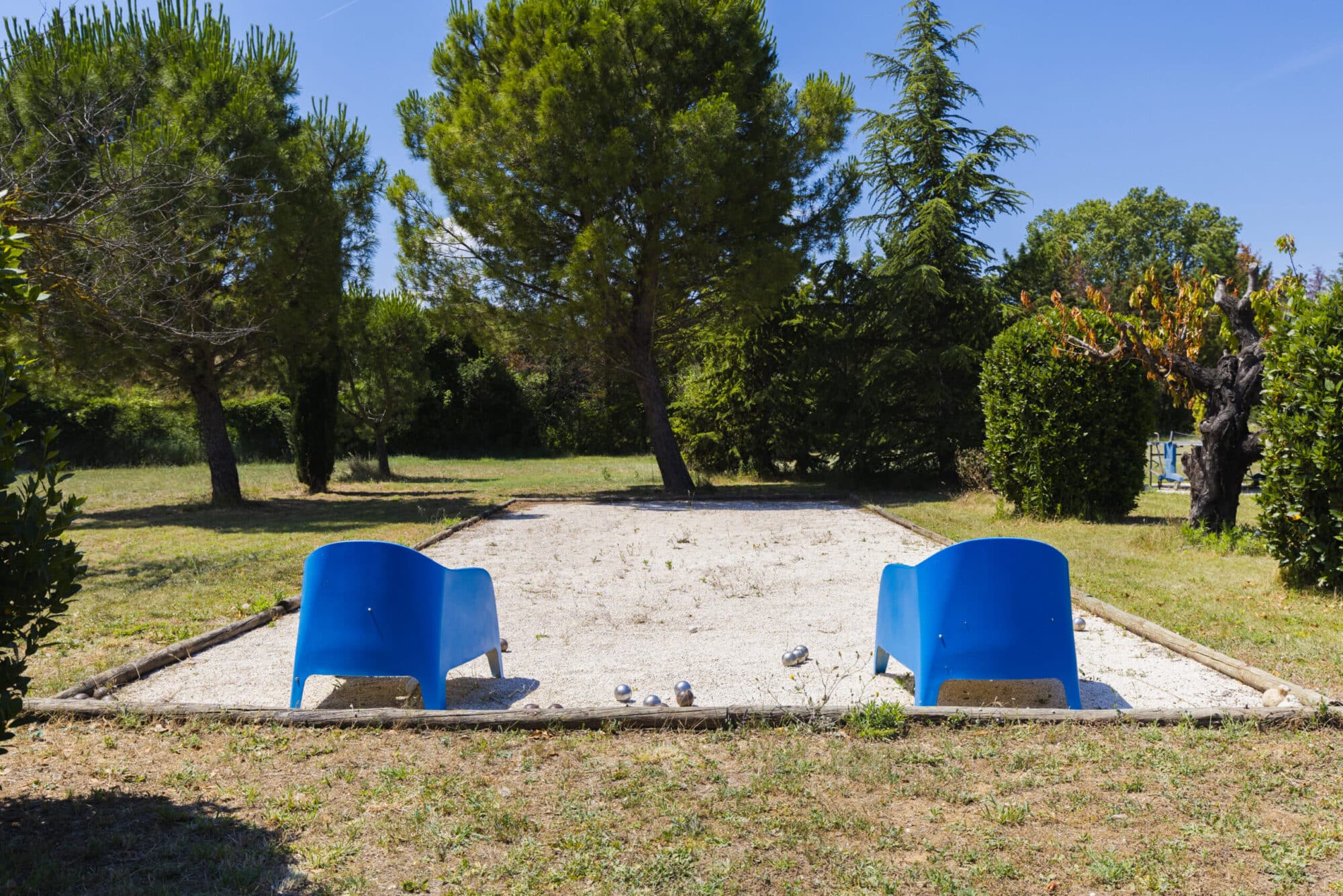 Terrain de pétanque en gravier avec fauteuils bleus, sous les pins du jardin