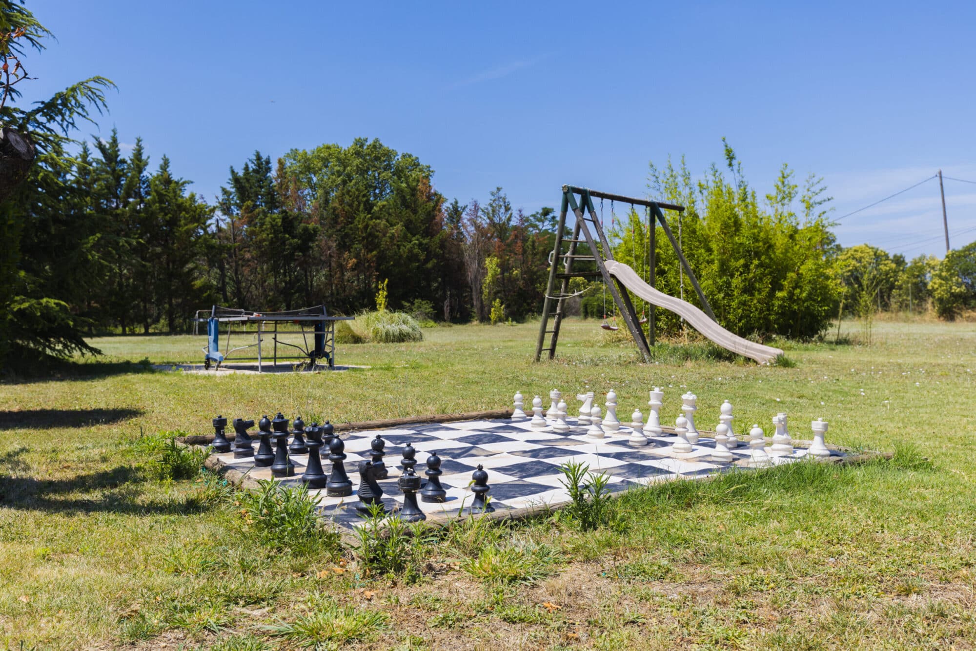 Échiquier géant avec pièces blanches et noires, devant une balançoire et un toboggan, dans le jardin